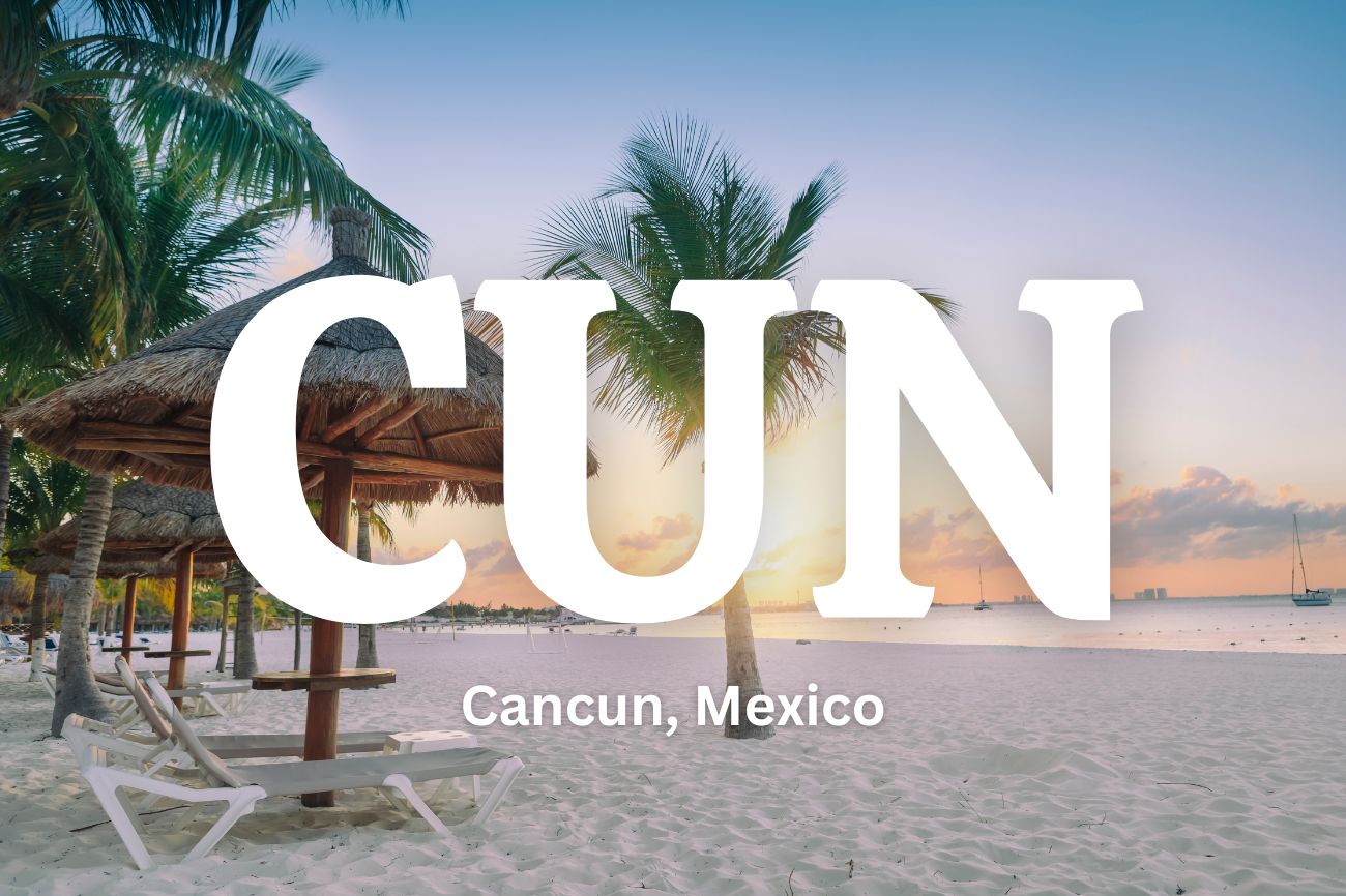 Text: CUN Cancun, Mexico on top of a picture of a sandy beach with a chair , umbrella and palm trees in the background at sunset